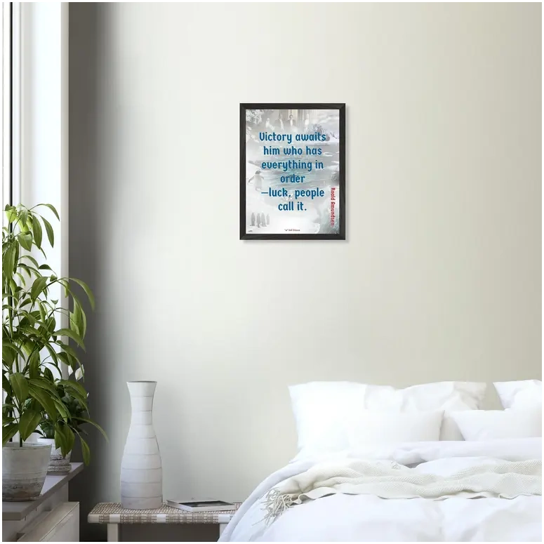 A black framed print with a quote from Roald Amundsen, 'Victory awaits him who has everything in order—luck people call it', hanging on a wall in a bedroom.