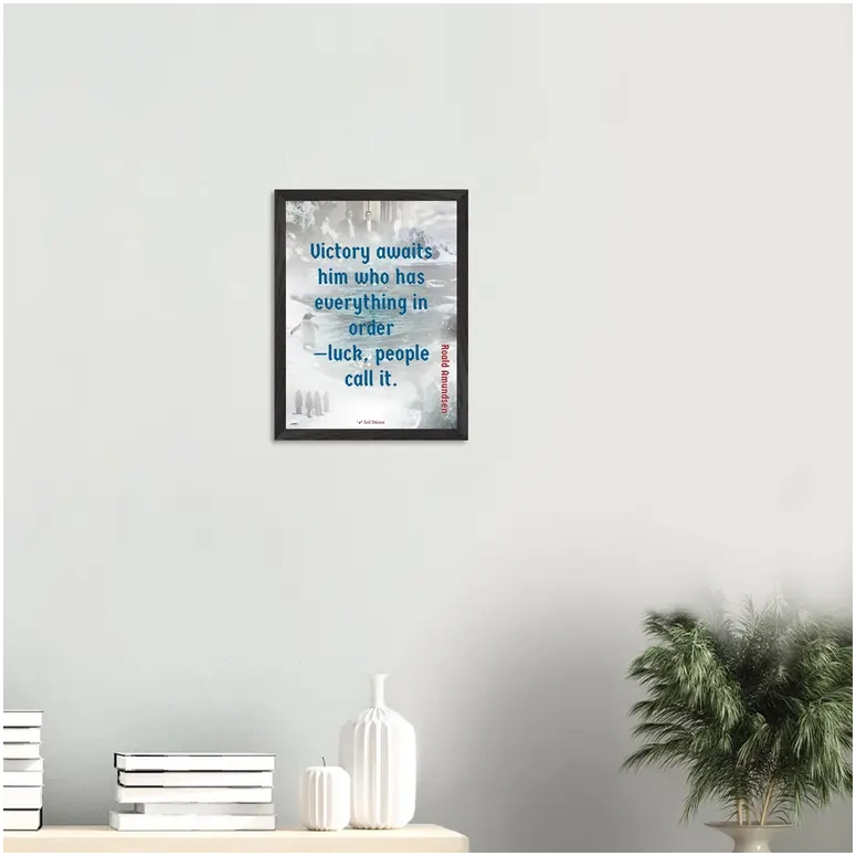 A black framed print with a quote from Roald Amundsen, 'Victory awaits him who has everything in order—luck people call it', hanging on a wall above a desk.